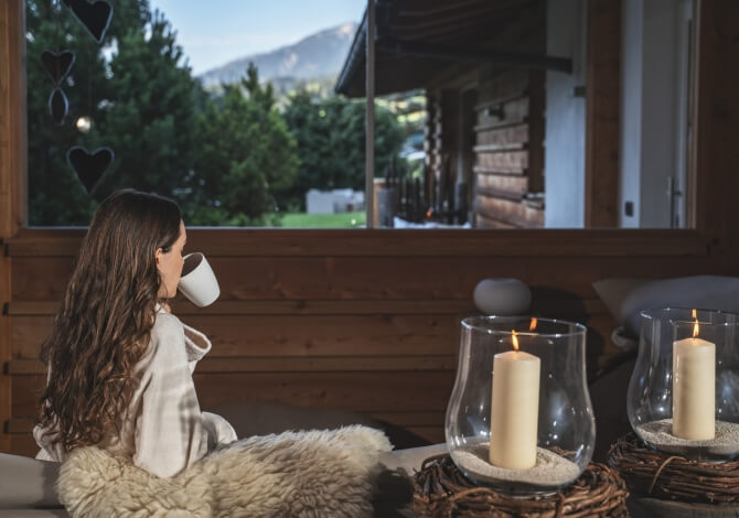 Una donna con capelli lunghi e rossi, in accappatoio bianco, sorseggia una tazza mentre guarda un paesaggio sereno dalla finestra.