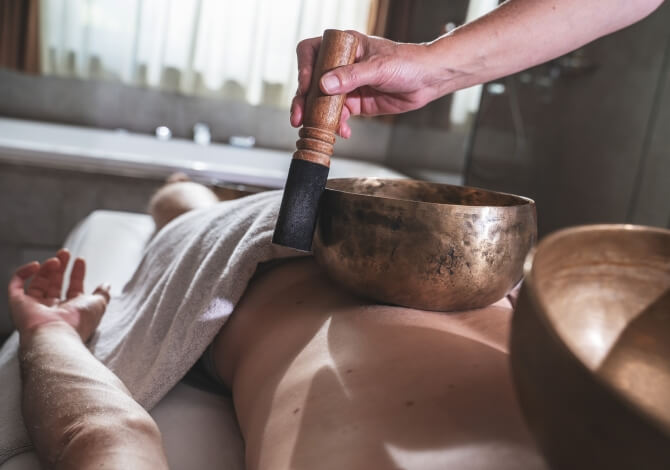 Una persona sdraiata con un asciugamano bianco, mentre un'altra tiene un martello sopra una ciotola in una stanza luminosa.