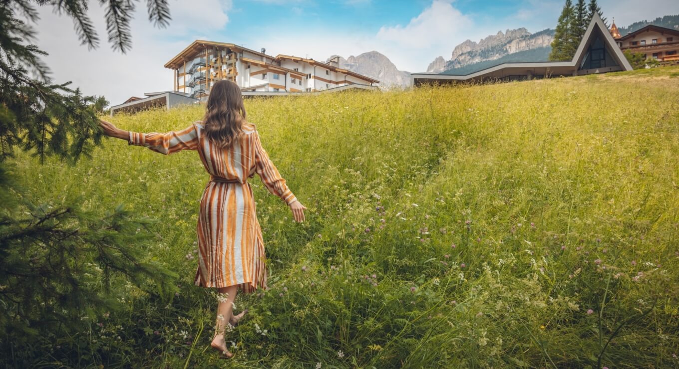 Una donna con un vestito a righe cammina a piedi nudi in un campo verde, con edifici e montagne sullo sfondo.