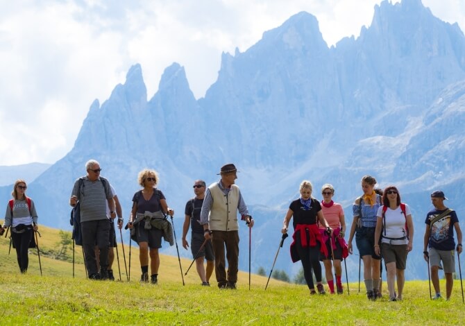 Gruppo di 12 persone in cammino nella campagna, circondato da montagne e cielo sereno, con bastoncini da trekking.
