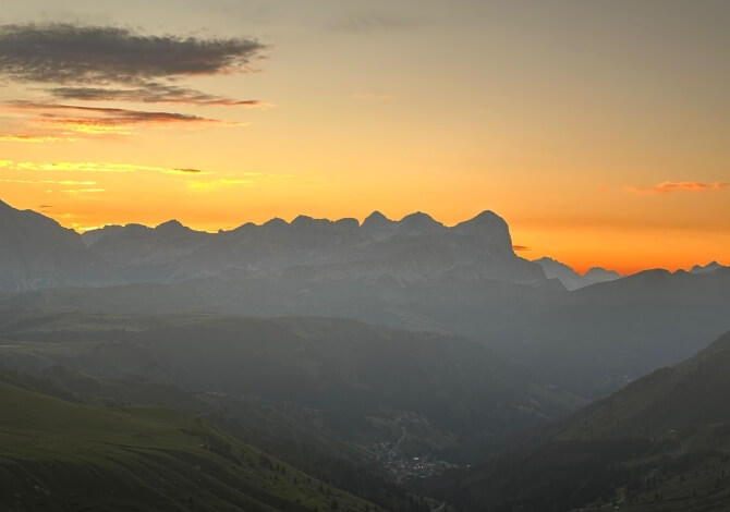Paesaggio montano sereno al tramonto, con cielo arancione e giallo, colline verdi e nebbia che avvolge le montagne.