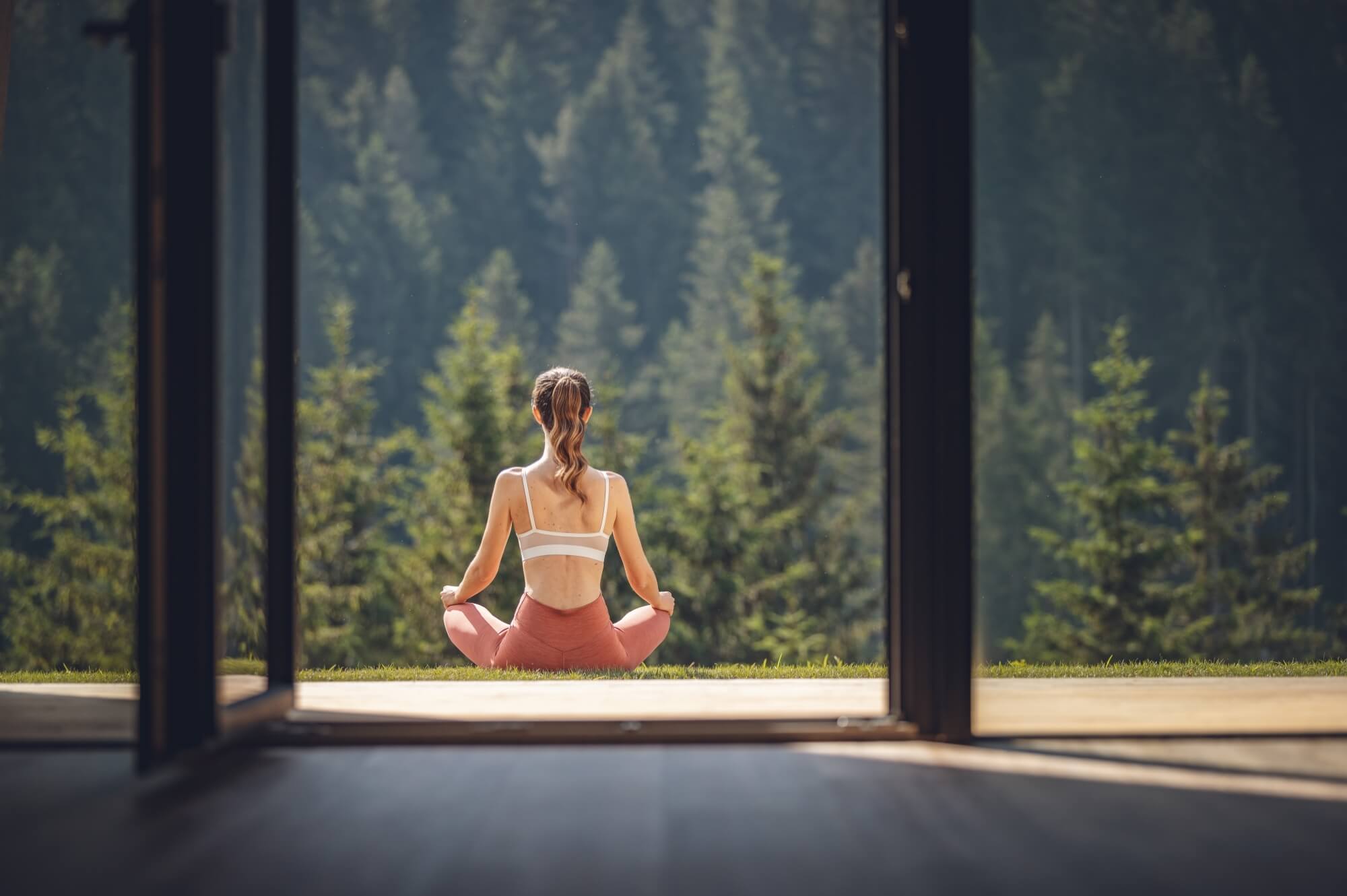 Donna che pratica yoga seduta davanti a una finestra, circondata da un paesaggio naturale sereno e tranquillo.