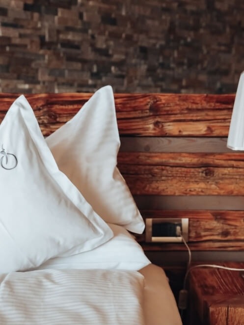 Letto con cuscini bianchi, testiera in legno, comodino con lampada e presa, su sfondo di muro in mattoni.