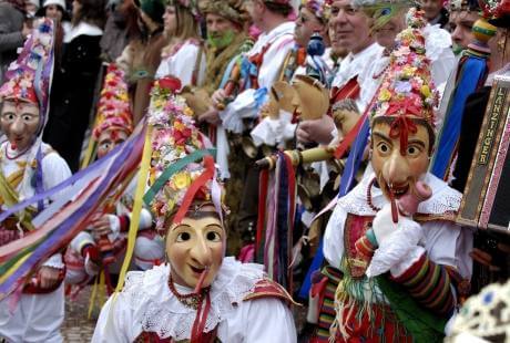 Gruppo di persone in abiti tradizionali con maschere, strumenti musicali e nastri colorati, durante un festival culturale.