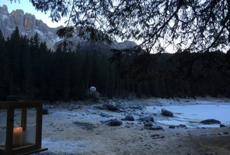 Paesaggio invernale sereno con lago ghiacciato, montagne innevate, alberi scuri e una lanterna che emette una luce calda.