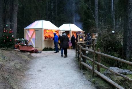 Scena invernale serena con due piccole capanne illuminate, un albero di Natale e persone che camminano lungo un sentiero.