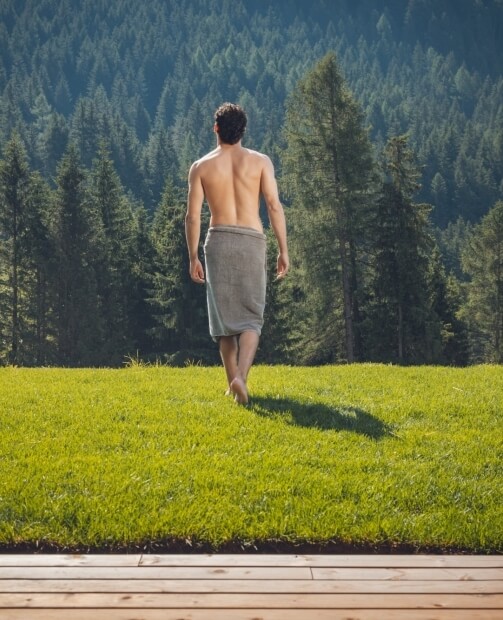Un uomo shirtless con un asciugamano in vita cammina su un ponte di legno verso una foresta, in un campo erboso.