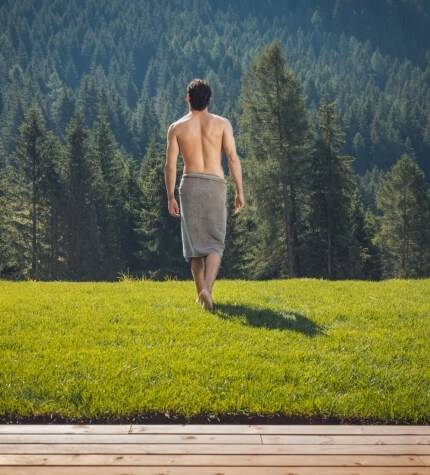 Un uomo con un asciugamano in vita cammina su un prato verde, con alberi e un ponte di legno sullo sfondo.