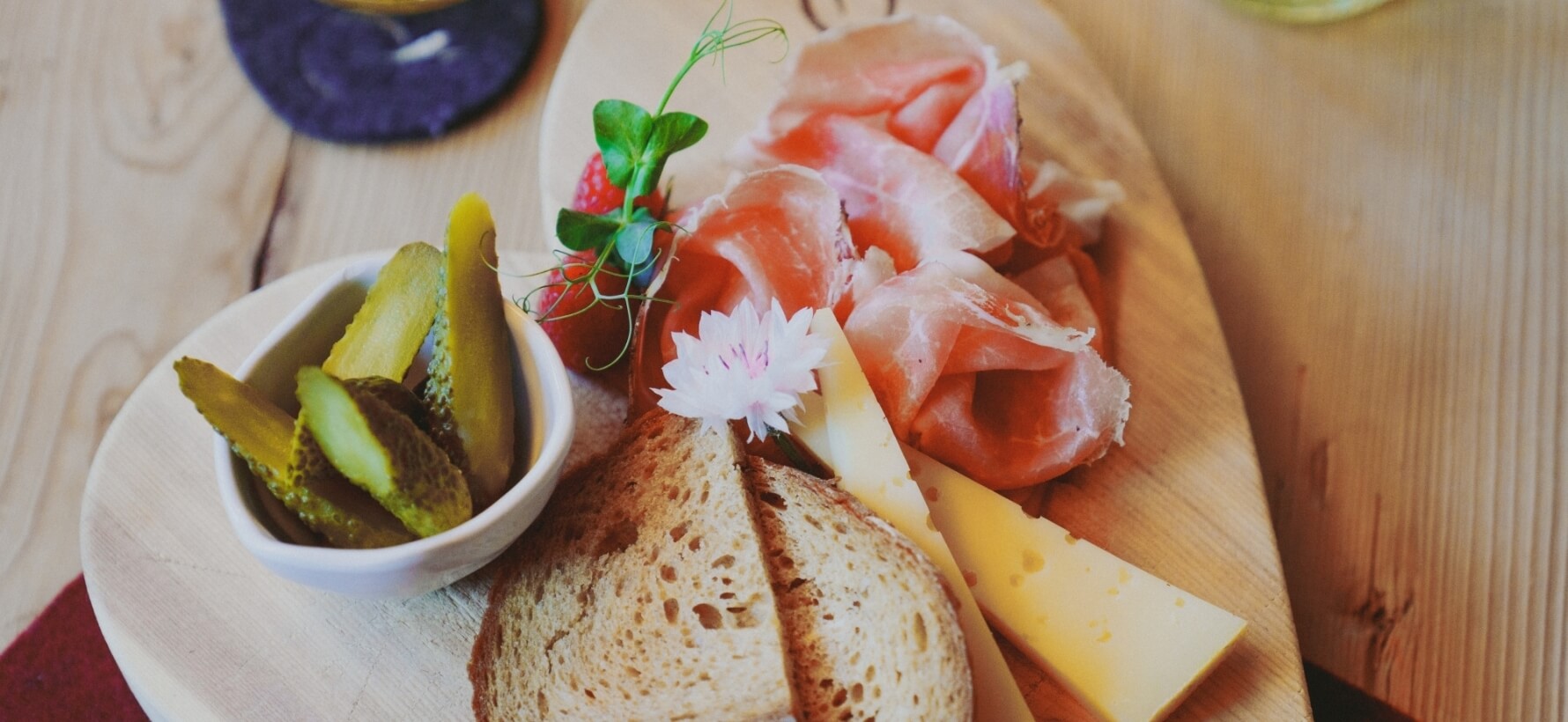 Disposizione di cibo su un tavolo di legno: piatto con formaggio, prosciutto, pane, cetrioli, fragola e fiore rosa.