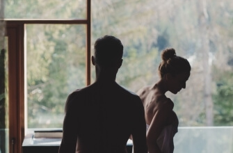 Uomo e donna shirtless davanti a una finestra, immersi in un'atmosfera intima e contemplativa, circondati da alberi.