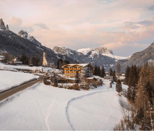 Scena invernale serena con paesaggio innevato, alberi, edifici giallo e bianco, catena montuosa e cielo nuvoloso rosa-grig