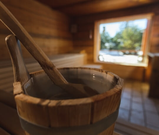 "Scena di sauna serena con secchio e mestolo in legno in primo piano, panche e finestra con vista sulla natura sullo sfondo."