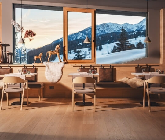 "Interno di un ristorante con panorama montuoso fuori dalle finestre, arredato con tavoli, sedie e una panca con tappeto di pelliccia