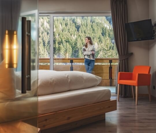 Camera d'albergo serena con vista su alberi, letto, sedia arancione e TV a schermo piatto; donna fuori dalla finestra.