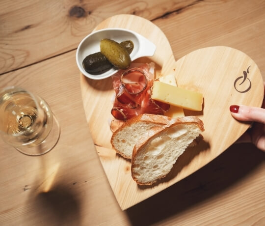 "Tavolo in legno con piatto a forma di cuore contenente pane, formaggio e sottaceti, accompagnato da un bicchiere di vino."