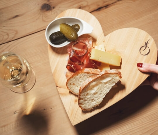 Tagliere di legno a forma di cuore con pane, formaggio, prosciutto, cetrioli e un bicchiere di vino bianco.