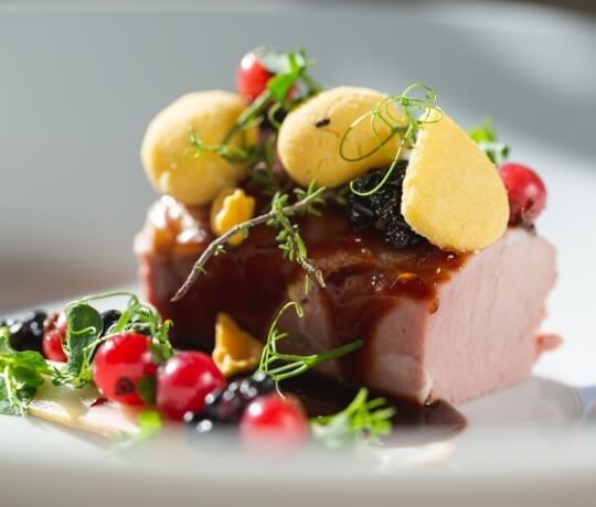 Fetta di prosciutto con salsa marrone, bacche rosse e guarnizioni nere su un piatto elegante con sfondo grigio chiaro.