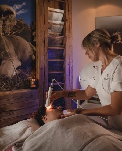 Una donna riceve un trattamento facciale mentre una terapista usa un dispositivo a luce rossa in una stanza bianca.