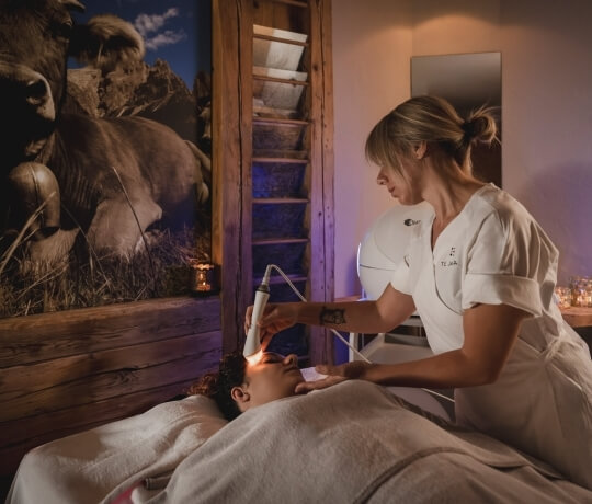 Una donna in un centro benessere esegue un trattamento facciale su un cliente sdraiato, con un murale di mucche sullo sfondo.