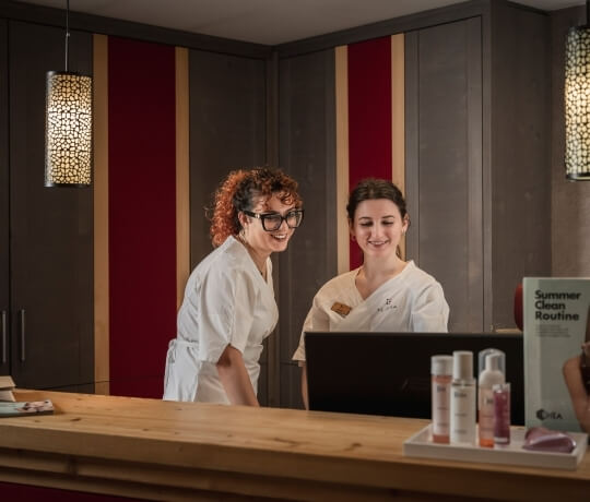 Due donne in divise bianche sorridono dietro un bancone di spa, guardando un laptop con prodotti di bellezza in vista.