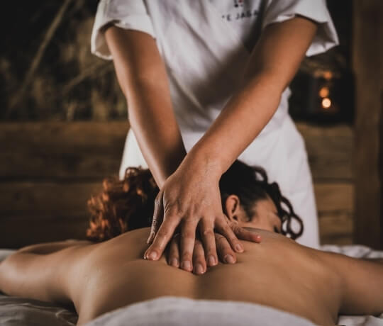 Due mani massaggiano la schiena di una persona sdraiata, in un ambiente intimo con luce soffusa e sfondo in legno scuro.