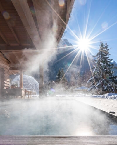 Una scena invernale serena con una vasca idromassaggio fumante su un deck di legno, circondata da alberi innevati.