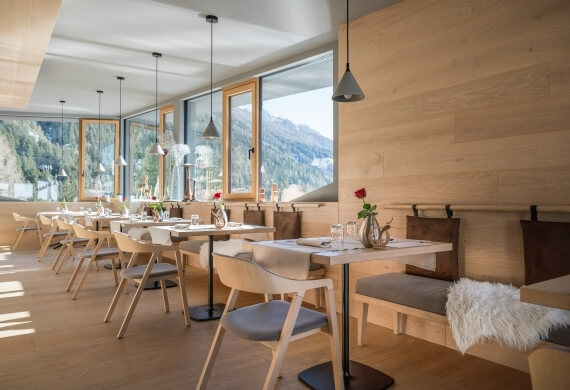 Area ristorante con tavoli e sedie, grande finestra con vista montagna, illuminazione soffusa e decorazioni sui tavoli.