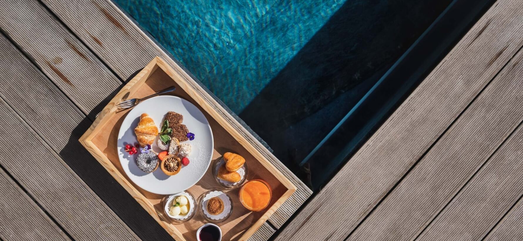 Vassoio di legno con dolci, ciotole di burro e succo, e bicchieri, accanto a una piscina blu.