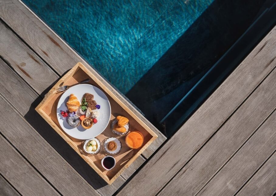 Vassoio per colazione su un deck con vista su una piscina, con dolci, frutta e bevande, in un'atmosfera accogliente.