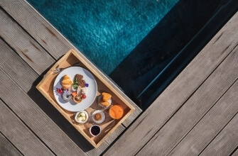 Vassoio di legno con colazione su un bordo piscina, con piatti colorati e bicchieri, su un decking in legno.