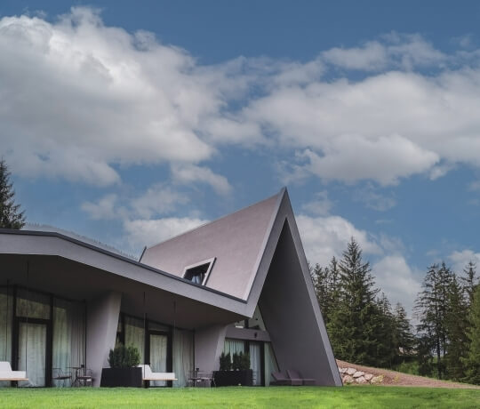 Retro di una casa moderna a due piani, con patio, giardino e alberi, sotto un cielo blu con nuvole bianche.