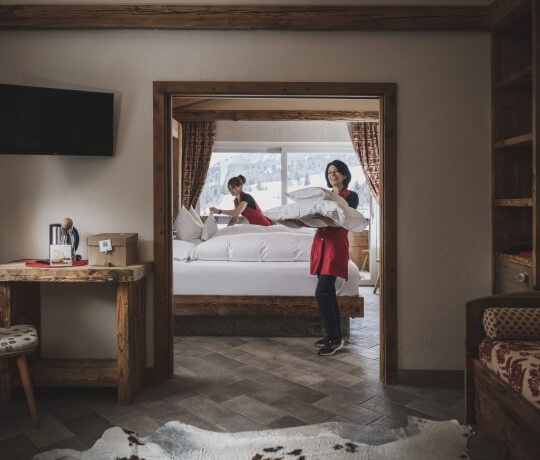 Due donne in un'accogliente stanza rustica, una con un vassoio e l'altra che rifà il letto, trasmettono serenità.