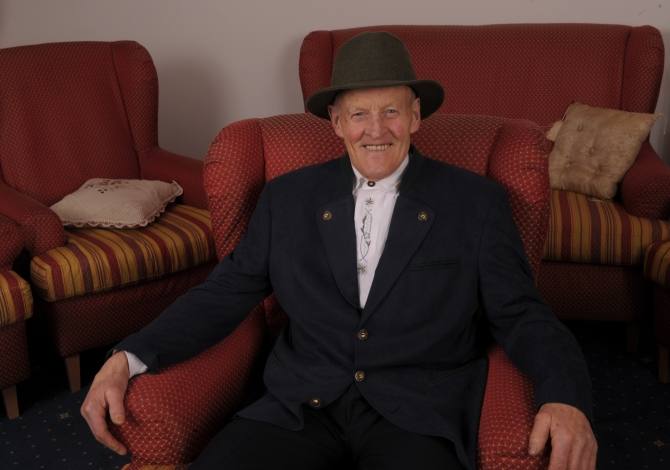 Un uomo anziano in un abito scuro e cappello, seduto su una poltrona rossa, circondato da sedie con cuscini a righe.