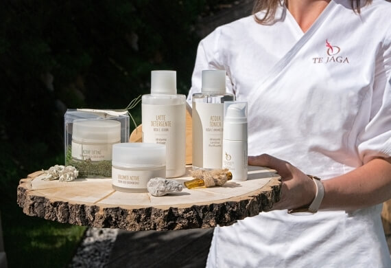 Donna con una maglietta "TEJAGA" che tiene un vassoio di legno con prodotti per la cura della pelle all'aperto.