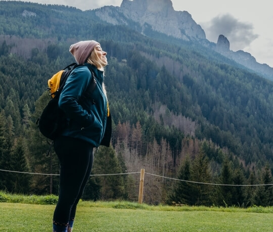 Donna con berretto rosa e zaino, montagna boscosa sullo sfondo.