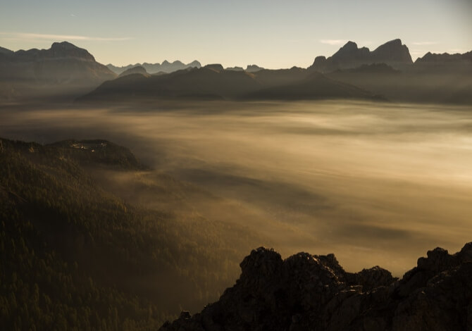 Paesaggio montano all'alba, con valli nebbiose illuminate da una luce dorata e montagne scure in primo piano.