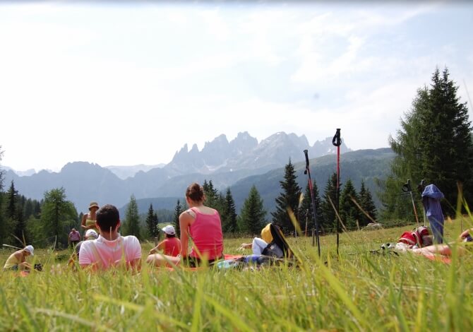 Persone si riuniscono in un campo erboso, con montagne maestose sullo sfondo e alberi ai lati.