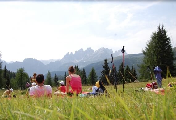 Persone sedute in un campo erboso, circondate da alberi e montagne, in un'atmosfera di relax e tranquillità.
