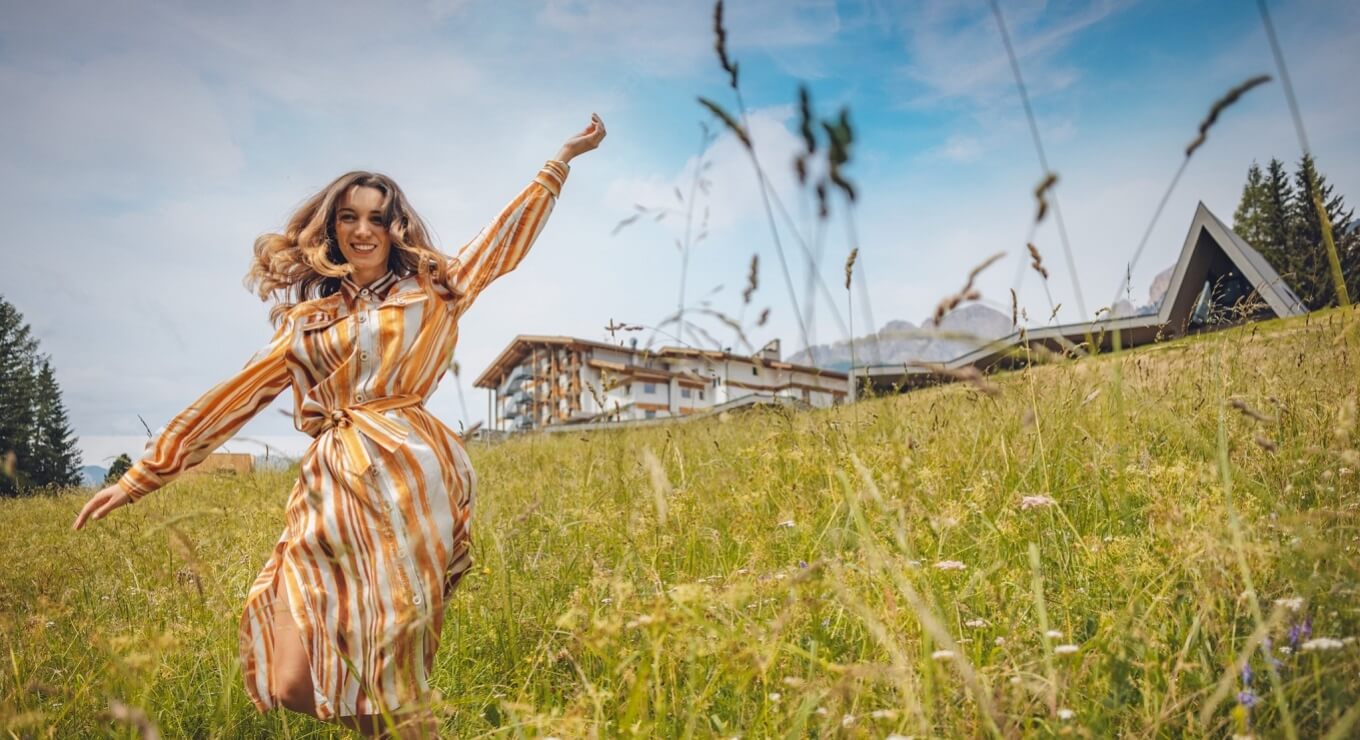 Una donna in un vestito arancione e bianco corre felice in un campo di erba alta e fiori selvatici, con un edificio A-frame sullo sfondo.