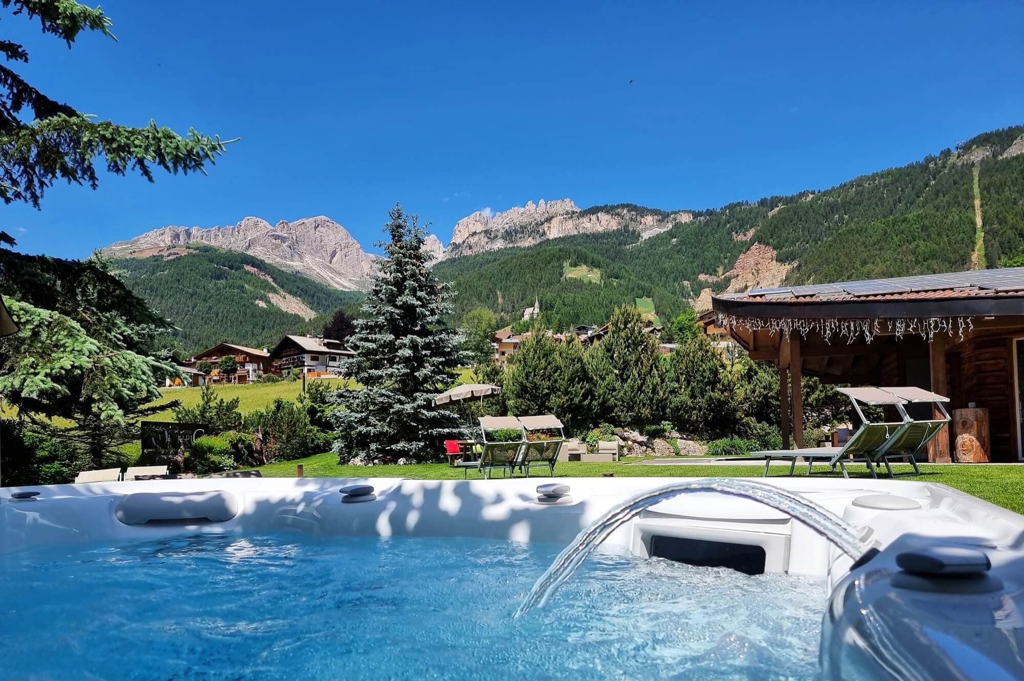 Una vasca idromassaggio in un giardino con montagne sullo sfondo, circondata da un patio e alberi verdi.