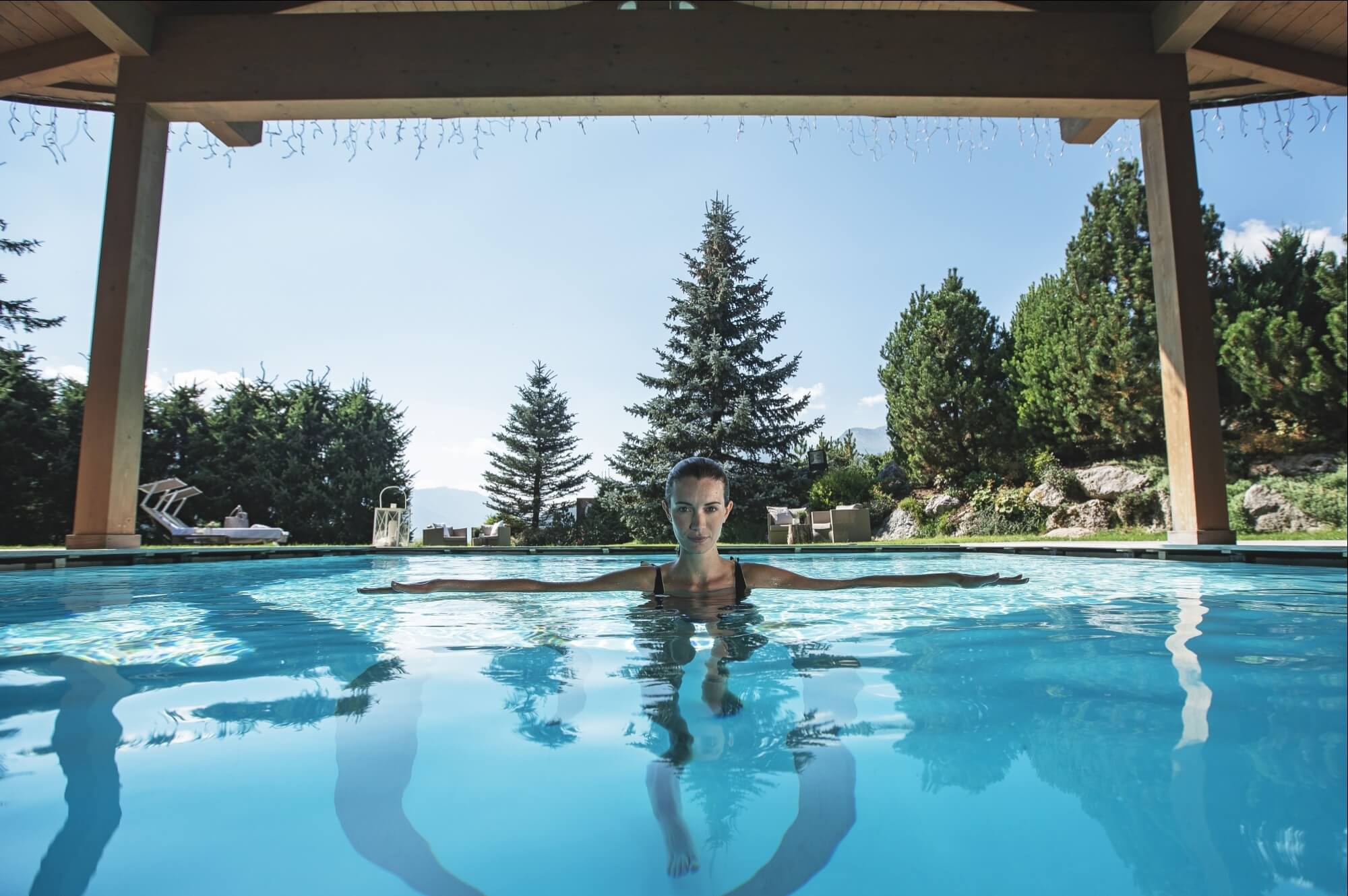 Donna che galleggia in una piscina circondata da alberi, con un cielo blu e luci appese sopra, atmosfera di relax.