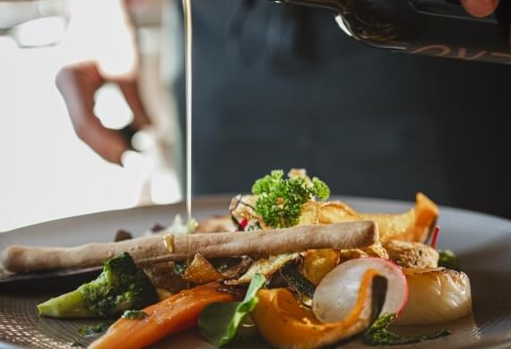 Primo piano di un piatto con verdure, pesce e un condimento versato da una bottiglia con "ENO" in un ristorante.