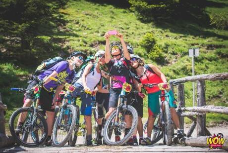 Un gruppo di sei ciclisti in mountain bike, in posa con oggetti rosa, davanti a un cancello di legno in un bosco.