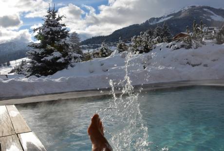 Una scena invernale serena con gambe immerse in acqua blu, circondate da alberi innevati e montagne.