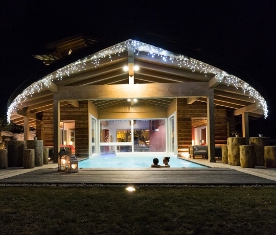 Struttura all'aperto con tetto a capanna, luci natalizie, piscina e due persone immerse, atmosfera di lusso e relax.