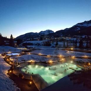 Una piscina all'aperto illuminata circondata da montagne innevate, con persone che si rilassano nell'acqua blu-verde.