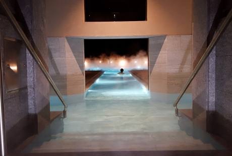 Una persona galleggia in una piscina serena con sfumature di blu, circondata da pareti in pietra chiara e scale in acciaio.