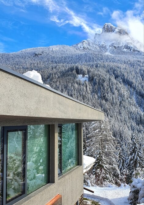 Edificio grigio moderno con finestre nere, davanti a una catena montuosa innevata e cielo blu, in un'atmosfera invernale.