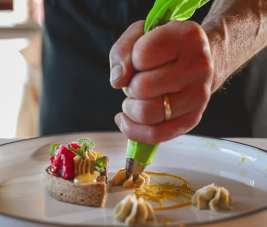 Una persona decora un tart con crema gialla e frutta rossa, usando una sac à poche verde su un piatto bianco.