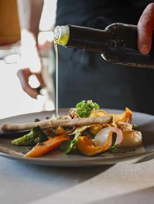 Una persona versa olio d'oliva su un piatto di verdure e pane, in un ambiente luminoso e curato.